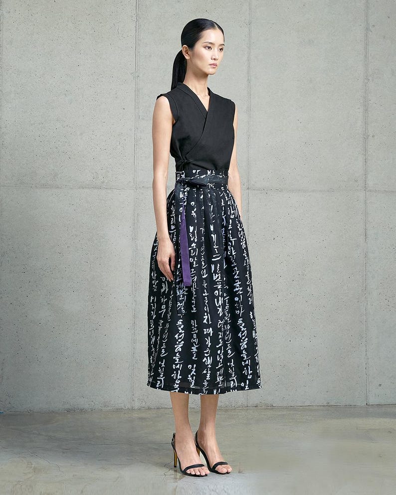 May include: A sleeveless black wrap top paired with a high-waisted midi skirt featuring a black and white calligraphic print. The skirt includes a wide waistband with a tie closure and a distinct purple fabric accent hanging from the side.