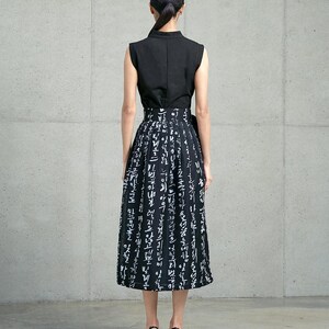 May include: A sleeveless black top paired with a high-waisted midi skirt featuring a black background and white Korean calligraphy print. The skirt has a pleated design and a side tie detail, worn with black heeled sandals.