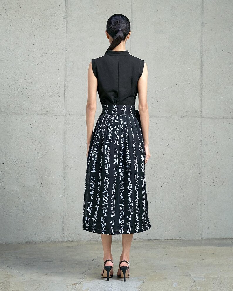 May include: A sleeveless black top paired with a high-waisted midi skirt featuring a black background and white Korean calligraphy print. The skirt has a pleated design and a side tie detail, worn with black heeled sandals.