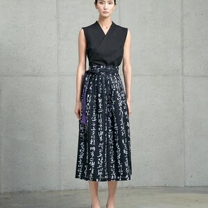 May include: A sleeveless black wrap top with a V-neckline paired with a black midi skirt featuring white Korean calligraphy print and a purple waist tie. The skirt has a structured waistband and falls below the knees in a straight silhouette.