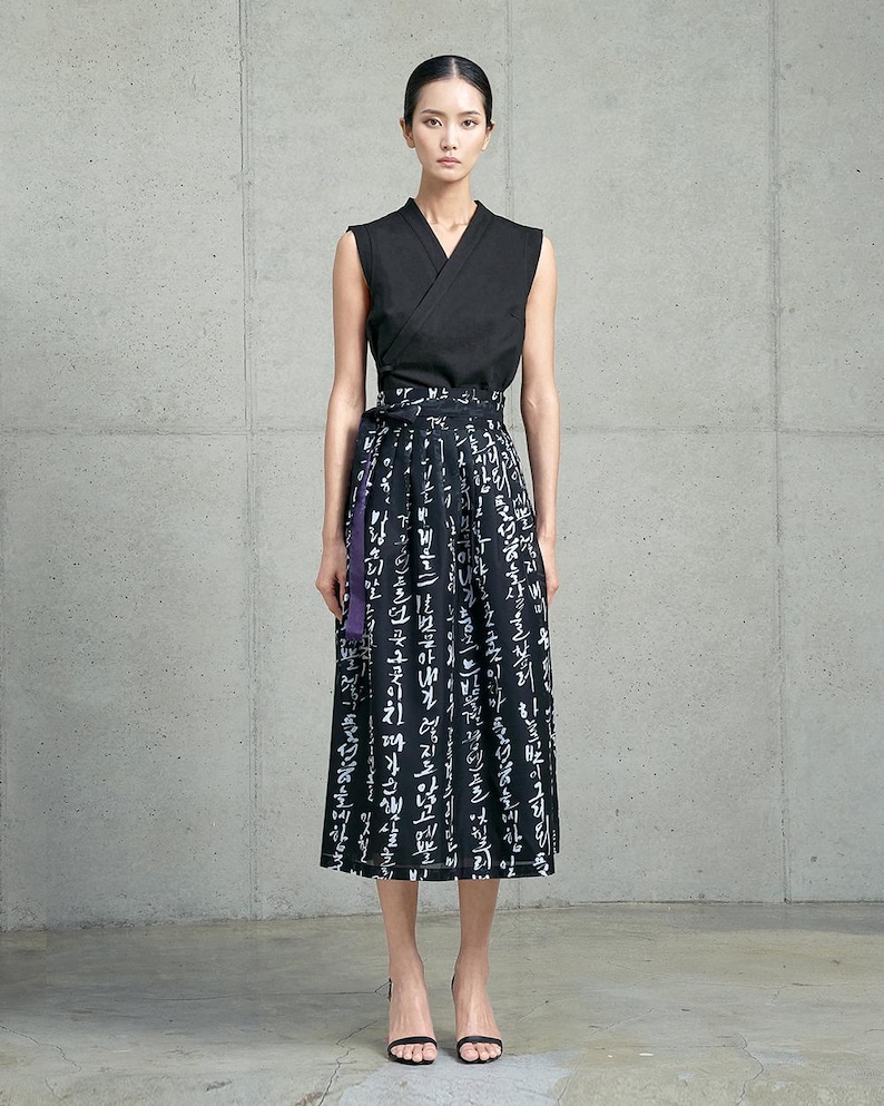 May include: A sleeveless black wrap top with a V-neckline paired with a black midi skirt featuring white Korean calligraphy print and a purple waist tie. The skirt has a structured waistband and falls below the knees in a straight silhouette.