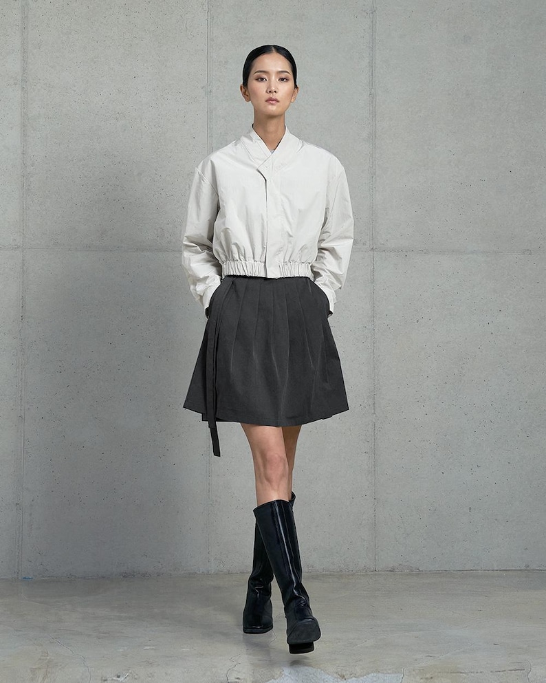 May include: A woman wearing a white collared shirt with a black skirt and black knee-high boots. The shirt has a cinched waist and the skirt has a pleated design.