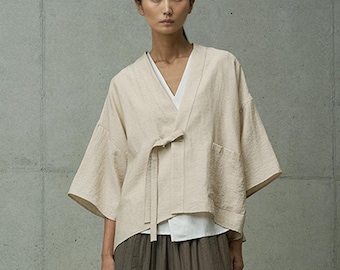 May include: A beige linen kimono-style jacket with a tie closure and a pocket on the left side. The jacket has a V-neck and is worn over a white shirt and brown pants.