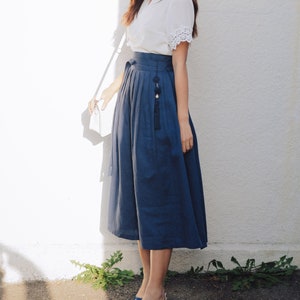 Puede incluir: Una mujer con una blusa blanca con detalles de encaje y una falda azul con un borla decorativa. Lleva tacones azules y lleva un bolso blanco.
