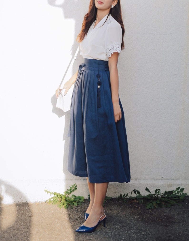 Puede incluir: Una mujer con una blusa blanca con detalles de encaje y una falda azul con un borla decorativa. Lleva tacones azules y lleva un bolso blanco.
