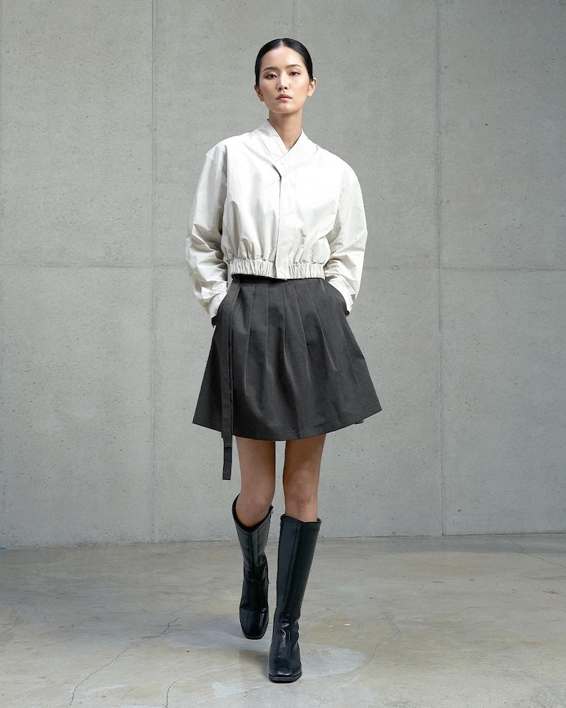 May include: A woman wearing a white, cropped, button-up shirt with a black skirt and black knee-high boots.