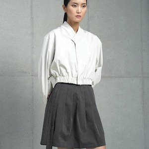May include: A woman is wearing a white, long-sleeved, collared shirt with a gathered waist and a dark gray, pleated skirt with a long tie.