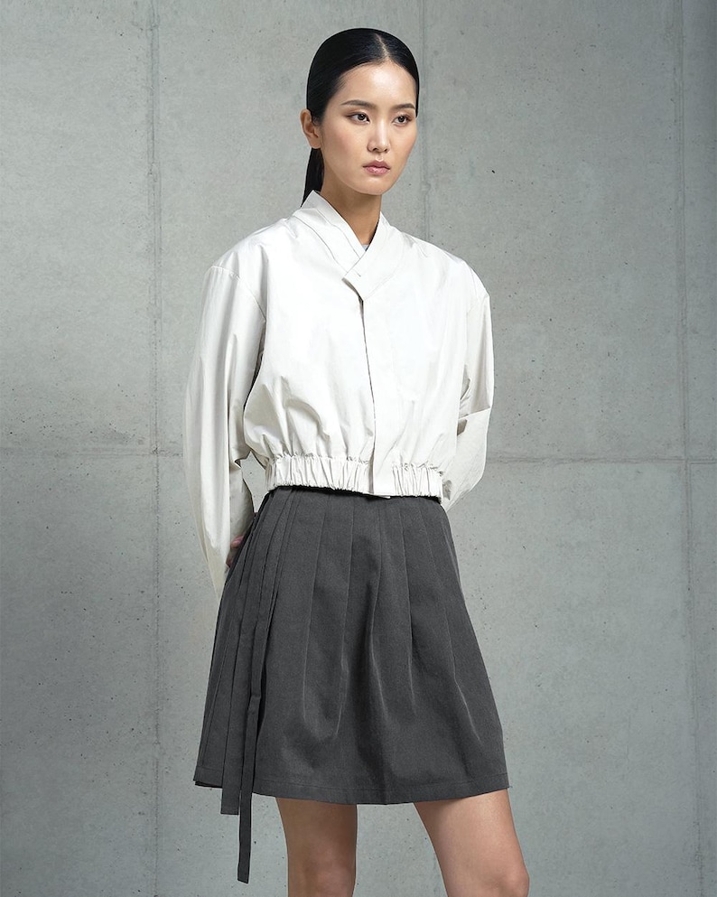 May include: A woman is wearing a white, long-sleeved, collared shirt with a gathered waist and a dark gray, pleated skirt with a long tie.