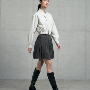May include: A woman wearing a white collared shirt with a black skirt and black knee-high boots. The skirt has a wrap design and a long tie.