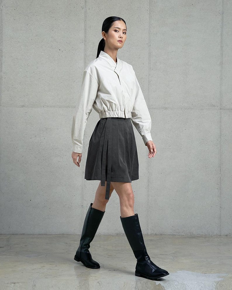 May include: A woman wearing a white collared shirt with a black skirt and black knee-high boots. The skirt has a wrap design and a long tie.