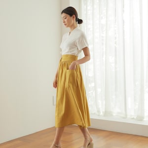 May include: A woman wearing a white short-sleeved blouse and a mustard yellow midi skirt with a pocket. She is wearing beige wedge sandals and standing on a wooden floor.