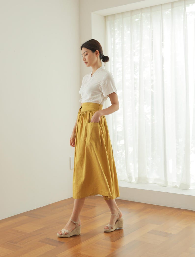 May include: A woman wearing a white short-sleeved blouse and a mustard yellow midi skirt with a pocket. She is wearing beige wedge sandals and standing on a wooden floor.