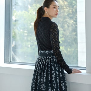 May include: A black long-sleeved top with a lace overlay and a high collar paired with a black skirt featuring a white printed pattern of Korean Hangul characters. The skirt has a wide waistband and a pleated design that falls to the lower legs.