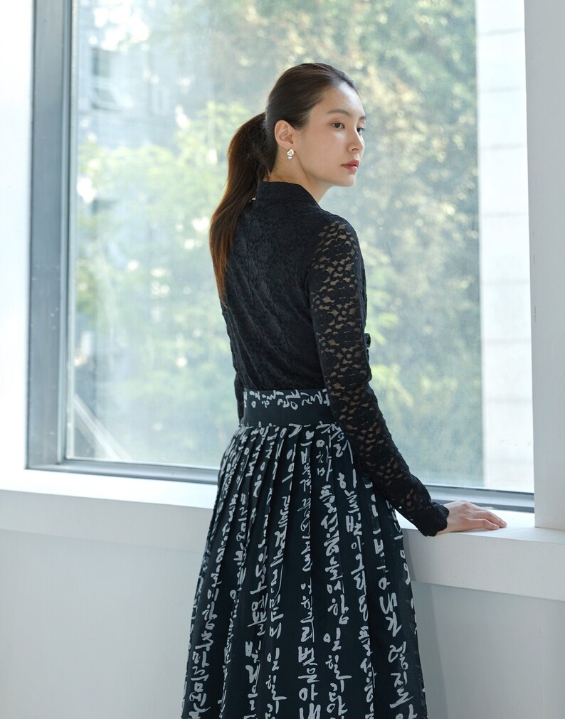 May include: A black long-sleeved top with a lace overlay and a high collar paired with a black skirt featuring a white printed pattern of Korean Hangul characters. The skirt has a wide waistband and a pleated design that falls to the lower legs.