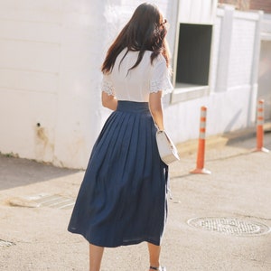 Puede incluir: Una mujer lleva una blusa blanca con ribetes de encaje y una falda plisada azul marino. Est&aacute; caminando por una calle y lleva un bolso blanco.