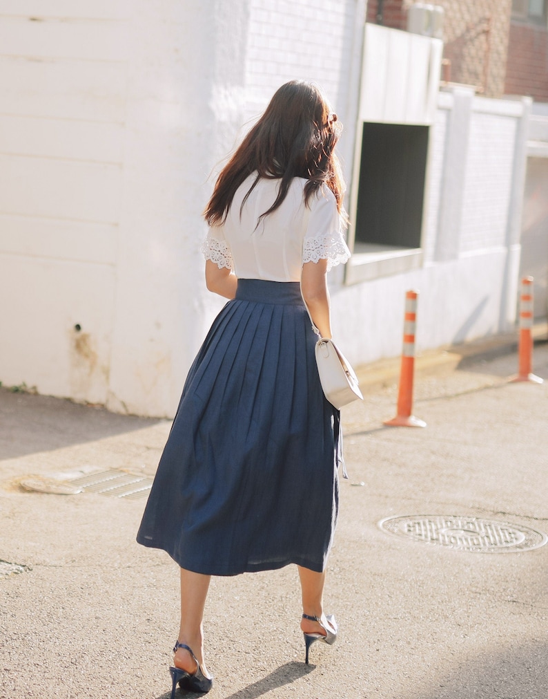 Puede incluir: Una mujer lleva una blusa blanca con ribetes de encaje y una falda plisada azul marino. Est&aacute; caminando por una calle y lleva un bolso blanco.