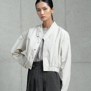 May include: A white cropped jacket with a V-neck and a drawstring waist. The jacket has a button closure and long sleeves. The model is wearing a gray shirt underneath the jacket and a black skirt.