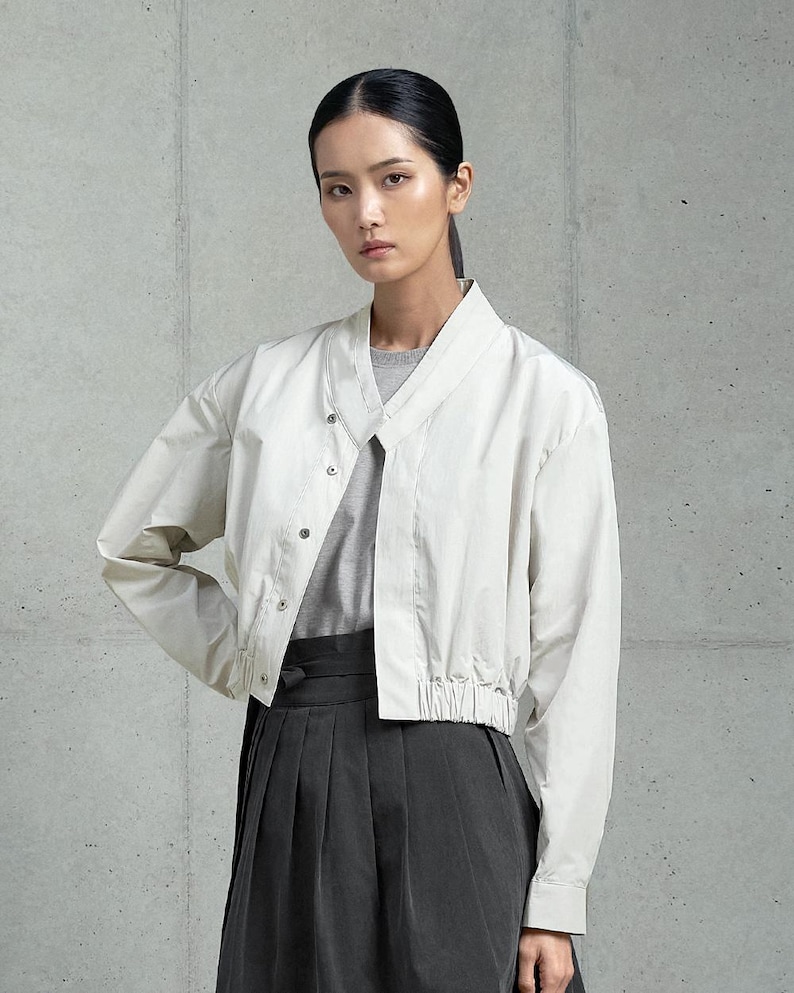 May include: A white cropped jacket with a V-neck and a drawstring waist. The jacket has a button closure and long sleeves. The model is wearing a gray shirt underneath the jacket and a black skirt.