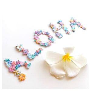 Könnte beinhalten: Das Wort "LOHA" in bunten, gestickten Blumenbuchstaben geschrieben. Eine weiße Frangipani-Blume mit gelbem Zentrum befindet sich in der unteren rechten Ecke. Die Buchstaben sind in Rosa-, Blau-, Orange- und Gelbtönen gehalten.