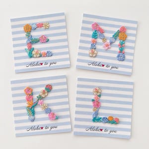 Può includere: Quattro biglietti quadrati con sfondi a righe blu e bianche. Ogni biglietto presenta una lettera ricamata diversa: E, M, K e L. Le lettere sono decorate con disegni floreali colorati. Il testo "Aloha to you" è stampato sotto ogni lettera.