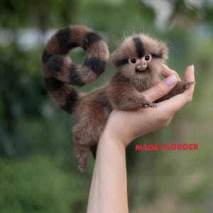 May include: A small, realistic plush monkey figurine with soft brown fur, a long striped tail curled into a circle, and dark facial markings. The figurine features large, expressive eyes and a small nose. The text MADE TO ORDER is visible.
