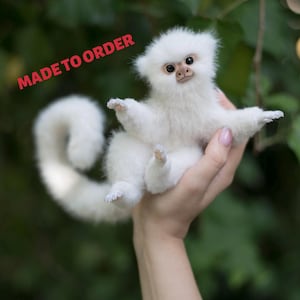 May include: A small, white, plush monkey figurine with a long, curved tail, dark eyes, and a light brown face. The text "MADE TO ORDER" is printed in red in the upper left corner. The figurine has soft, fluffy fur and detailed paws.