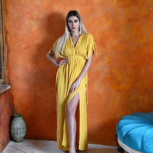 K&ouml;nnte beinhalten: Eine Frau tr&auml;gt ein langes, gelbes Maxikleid mit V-Ausschnitt und seitlichem Schlitz. Das Kleid hat eine taillierte Taille und kurze &Auml;rmel. Sie steht vor einer strukturierten orangen Wand.