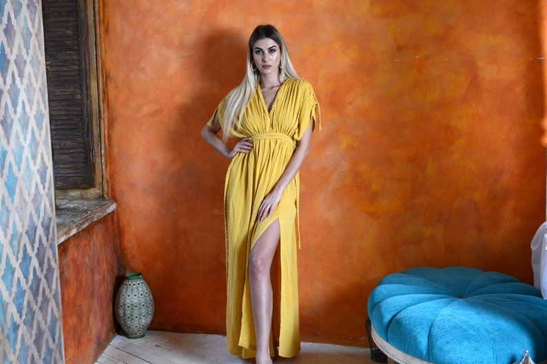 K&ouml;nnte beinhalten: Eine Frau tr&auml;gt ein langes, gelbes Maxikleid mit V-Ausschnitt und seitlichem Schlitz. Das Kleid hat eine taillierte Taille und kurze &Auml;rmel. Sie steht vor einer strukturierten orangen Wand.