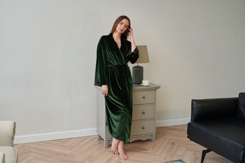 K&ouml;nnte beinhalten: Eine lange, dunkelgr&uuml;ne Samt-Robe mit Schalkragen und einem Bindeg&uuml;rtel. Die Robe wird von einer Person getragen, die in einem Raum mit Holzboden und wei&szlig;er Wand steht.