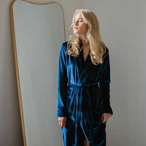 May include: A long, teal blue velvet robe with a matching tie belt. The robe has long sleeves and a deep V-neck. A large, gold-framed mirror is in the background.