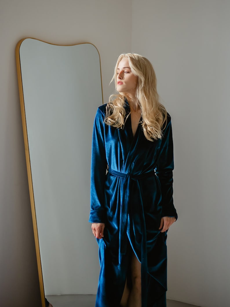 May include: A long, teal blue velvet robe with a matching tie belt. The robe has long sleeves and a deep V-neck. A large, gold-framed mirror is in the background.