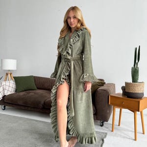 May include: A sage green velvet robe with ruffled trim and a matching belt. The long-sleeved robe features a cascading ruffle down the front and around the hem. The model is wearing the robe in a home setting.