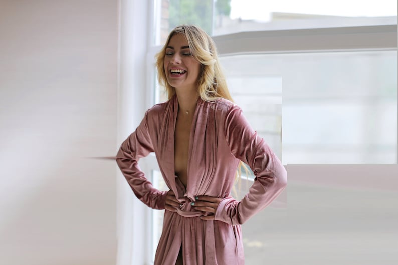 May include: A woman in a pink velvet robe with a deep V-neckline, standing in front of a window. She is smiling and looking to the side.