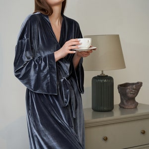 K&ouml;nnte beinhalten: Eine Frau in einem dunkelblauen Samt-Morgenmantel h&auml;lt eine wei&szlig;e Tasse und Untertasse. Sie steht vor einer Kommode mit einer Lampe und einer dekorativen Skulptur.