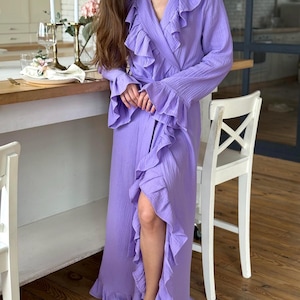 May include: A long, lavender-colored robe with ruffled trim and long sleeves. The robe has a wrap-around design and is made of a lightweight fabric. The model is standing in a kitchen setting.