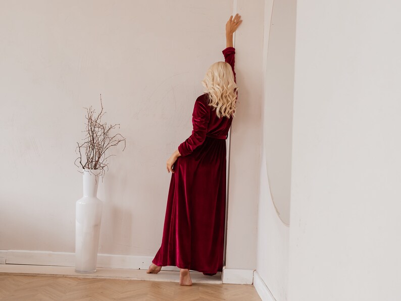 May include: A long, burgundy velvet robe with long sleeves. The robe is floor-length and has a wrap-around design. A tall white vase with branches is in the background.
