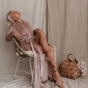 May include: A woman wearing a blush pink velvet robe sits in a wicker chair with her legs crossed. She is wearing a headband and has her eyes closed. A woven basket with flowers sits on the floor next to her.