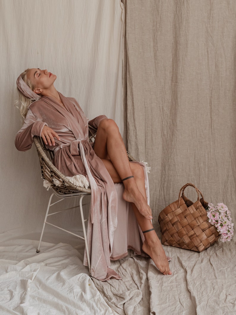 May include: A woman wearing a blush pink velvet robe sits in a wicker chair with her legs crossed. She is wearing a headband and has her eyes closed. A woven basket with flowers sits on the floor next to her.