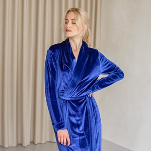 May include: A long, royal blue velvet robe with a shawl collar and a waist tie. The robe has long sleeves and is made of a soft, plush material. The model is wearing the robe in a studio setting.