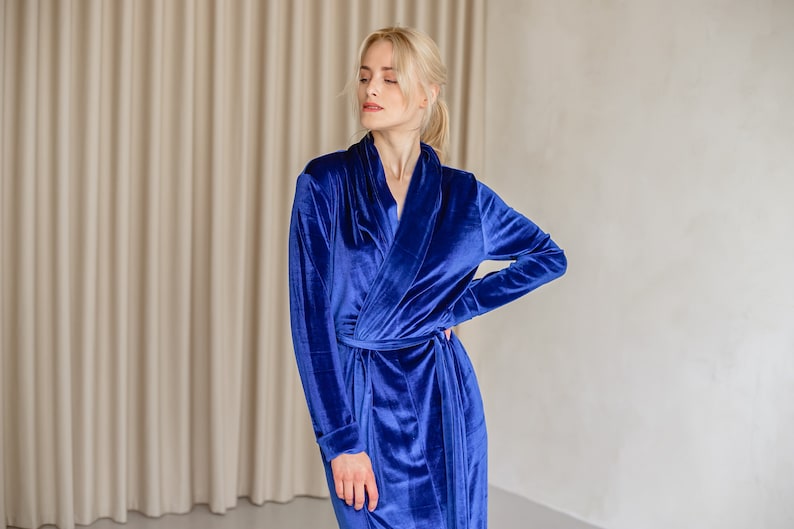 May include: A long, royal blue velvet robe with a shawl collar and a waist tie. The robe has long sleeves and is made of a soft, plush material. The model is wearing the robe in a studio setting.