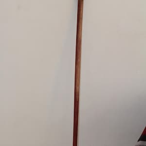 May include: A wooden walking cane with a dark brown handle and a long, slender shaft. The handle has a decorative, textured design. The cane is made of a light brown wood with a darker brown accent.