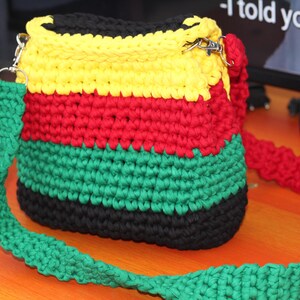May include: A colourful crochet bag in black, yellow, red, and green. The bag is rectangular with a long strap. The strap and the bag's body are made of the same crochet material. The bag has metal clasps.