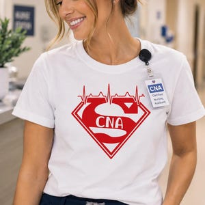 May include: A white short-sleeved t-shirt featuring a red graphic of a diamond-shaped emblem with a heartbeat line across the top and the letters CNA inside. The shirt is paired with dark navy blue scrub pants with a drawstring waistband.