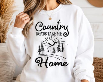 May include: A white crewneck sweatshirt featuring black screen-printed text that reads Country ROADS TAKE ME Home. The design includes a graphic of a sun rising over mountain peaks, pine trees, and a winding road leading toward the horizon.