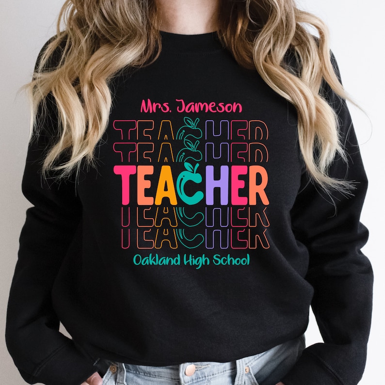May include: Black sweatshirt with a colorful "Teacher" graphic with an apple in the "O". The text "Mrs. Jameson" and "Oakland High School" are also printed on the sweatshirt.
