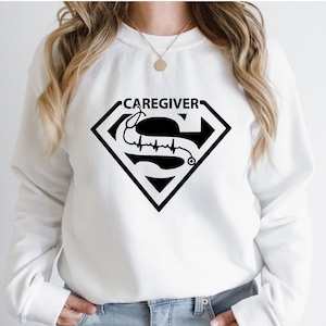 May include: White sweatshirt with a black Superman logo that says "Caregiver" with a stethoscope and a heartbeat line inside the logo.