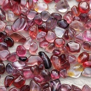 May include: A collection of pink, red, and purple tumbled stones. The stones are smooth and polished, and they have a variety of shapes and sizes.