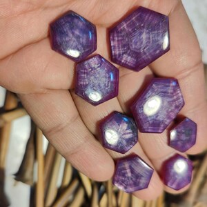 Puede incluir: Una colección de gemas hexagonales facetadas de varios tamaños. Las gemas son de color púrpura intenso con patrones internos que crean un efecto de estallido estelar. Las piedras se sostienen en una mano, mostrando sus superficies pulidas y diseños únicos.