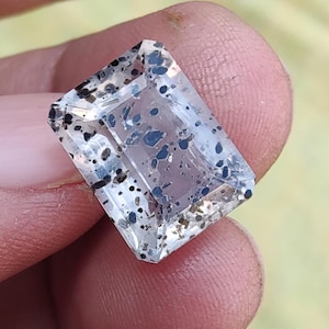 May include: A clear, rectangular gemstone with black and blue inclusions. The gem is held between fingers, showcasing its facets and internal patterns. The stone's clarity and the distribution of the dark spots are prominent.