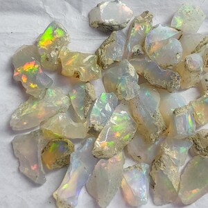 May include: A collection of raw opal gemstones. The opals display a milky white base color with flashes of iridescent colors, including green, yellow, and orange. The rough stones have a natural, unpolished appearance.